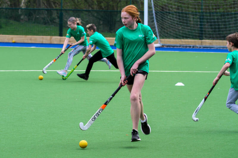 Group of children in green t-shirts playing hockey on an artificial pitch