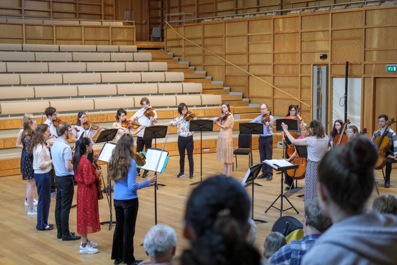 Group of string players standing performing in a concert hall, view from amongst the audience