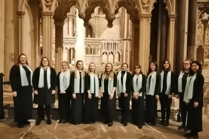 Row of singers wearing formal black with light blue scarves standing in front of some ornate-carved stonework