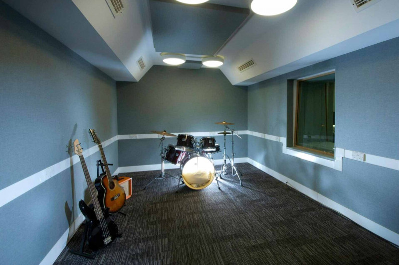 A room containing a drum kit and two guitars. The room has one internal window and bright ceiling lights.