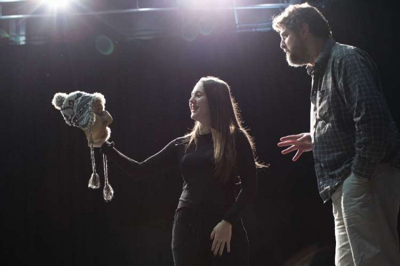 A female student in a dimly lit studio holds up a theatrical mask as her lecturer gives guidance.
