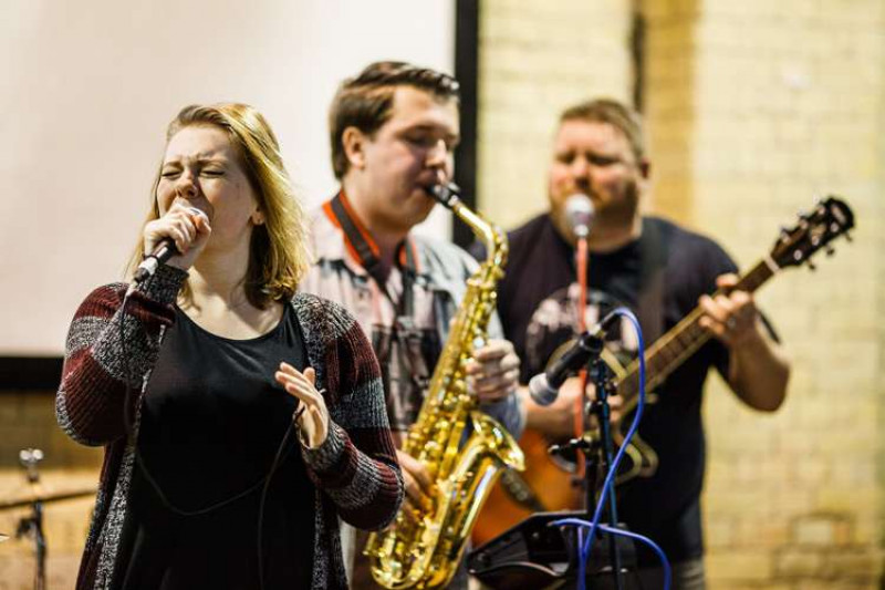 A female singer, a male saxophonist and a male guitar player perform at an indoor venue.