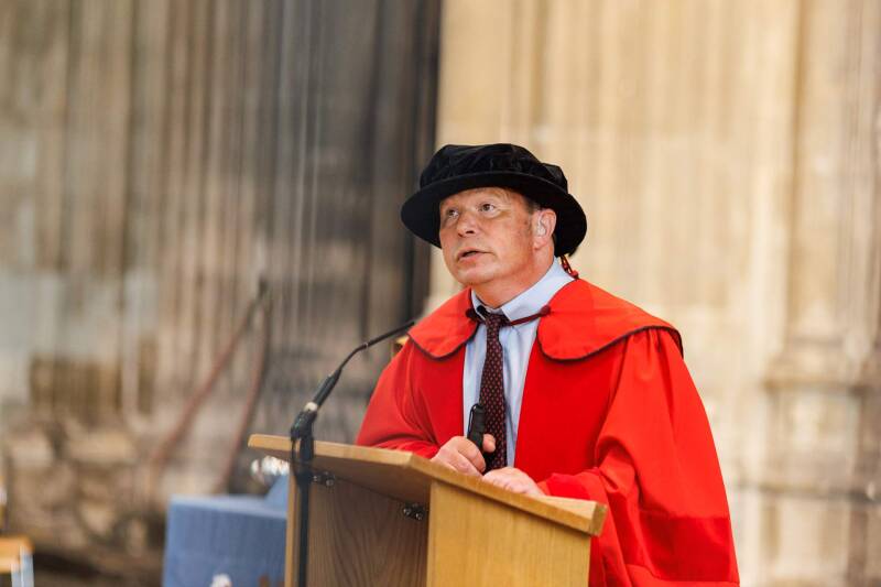 Howard Leicester speaking in Canterbury Cathedral at his honorary degree conferral, wearing red robes and a tudor bonnet