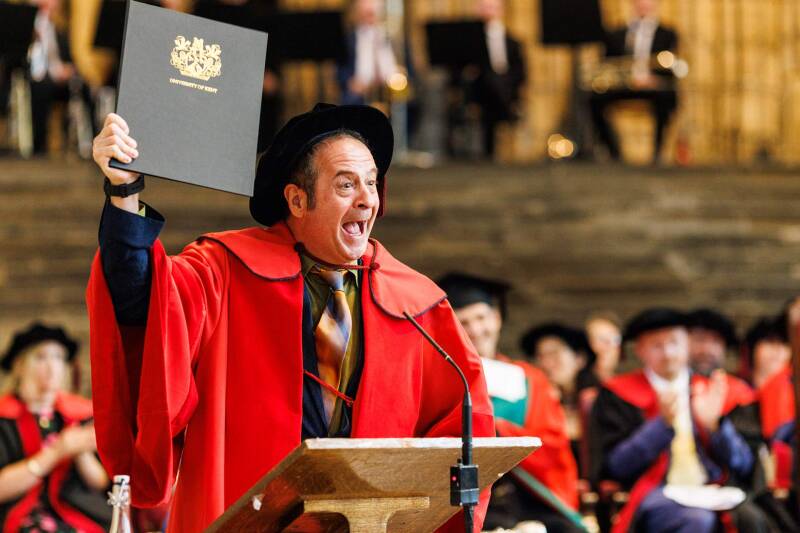 Mark Thomas speaking in Canterbury Cathedral, wearing red robes and a tudor bonnet, and brandishing his certificate folder