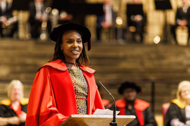 Olufunke Abimbola MBE speaking in Canterbury Cathedral at her honorary degree conferral, wearing red robes and a tudor bonnet