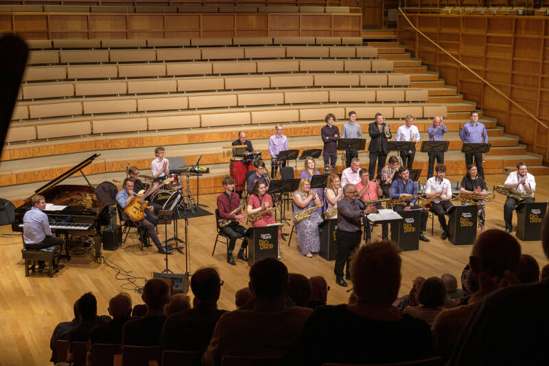 A group of jazz musicians listen as the conductor addresses the audience in a concert-hall