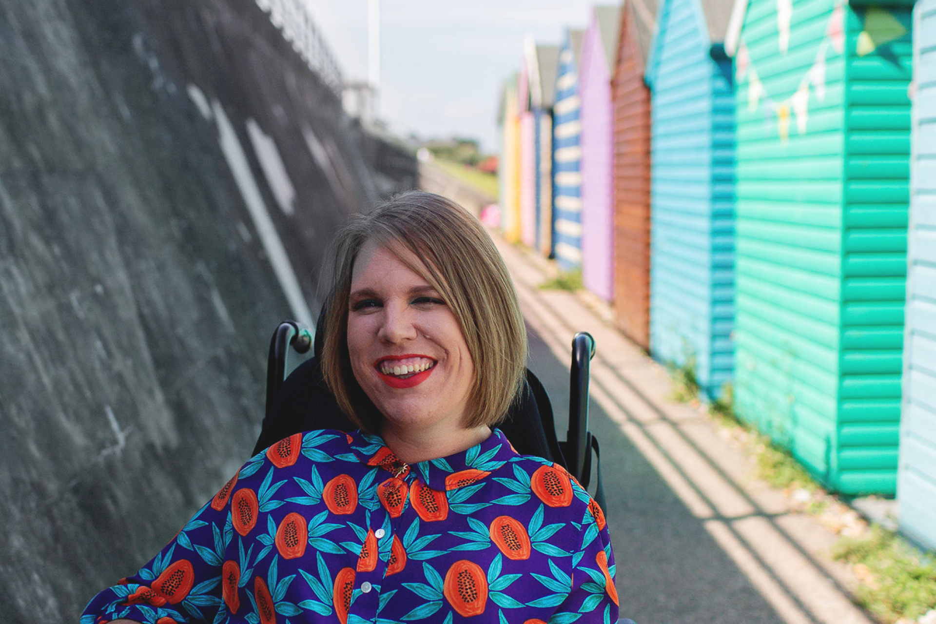Woman in wheelchair on promenade near brightly coloured beach huts.