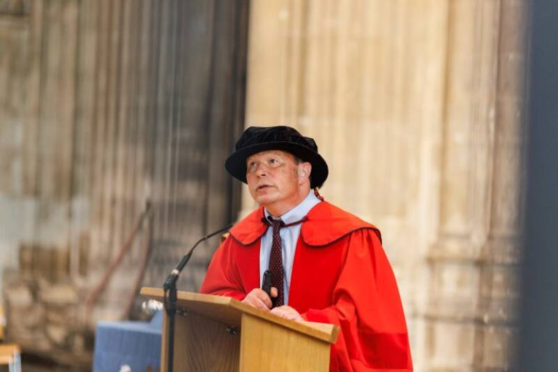 Man in graduation robes speaking at lectern in a cathedral.