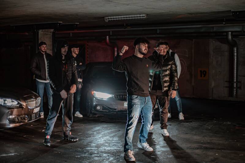 A dark underground car park. Six men stand in front of two BMW cars. The man at the front has his fists raised.  S