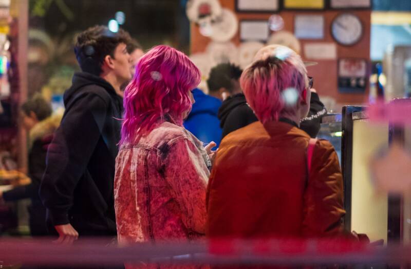Two young women with pink hair seen from the back standing in a busy chip shop at night.