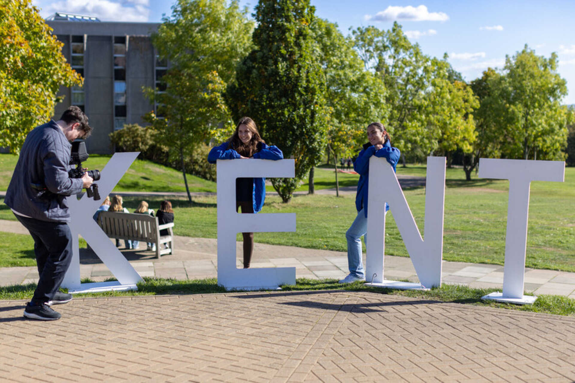 Two students being filmed standing by a large letters spelling 'Kent'