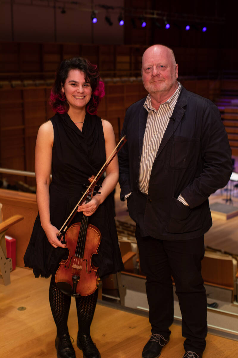 Female viola player and male composer standing on a balcony at the back of a concert hall