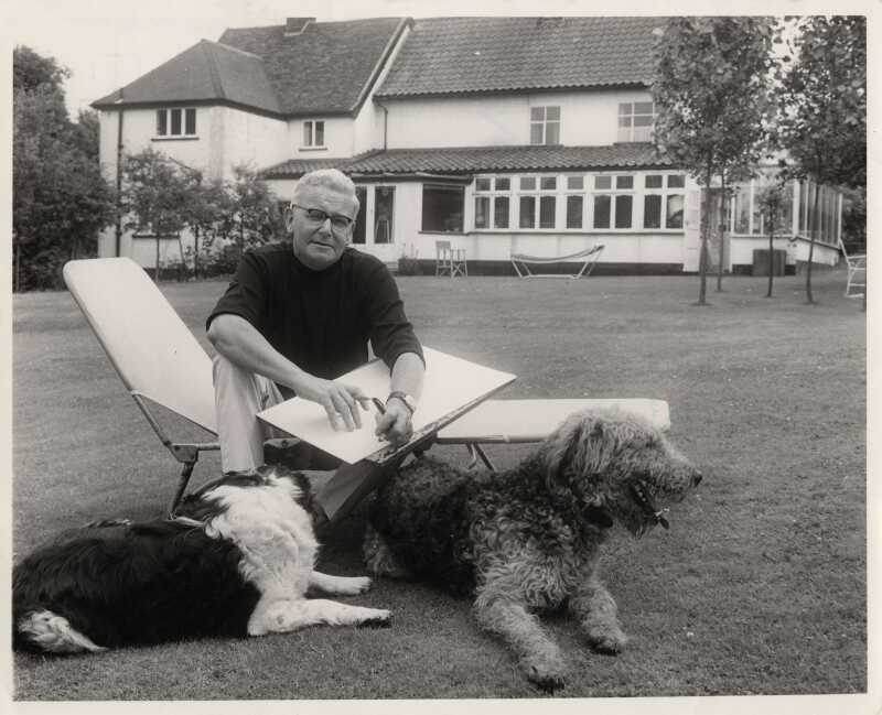 Black and white photo of Giles sitting in the back garden of Hillbrow Farm, holding sketch and drawing board, with his dogs