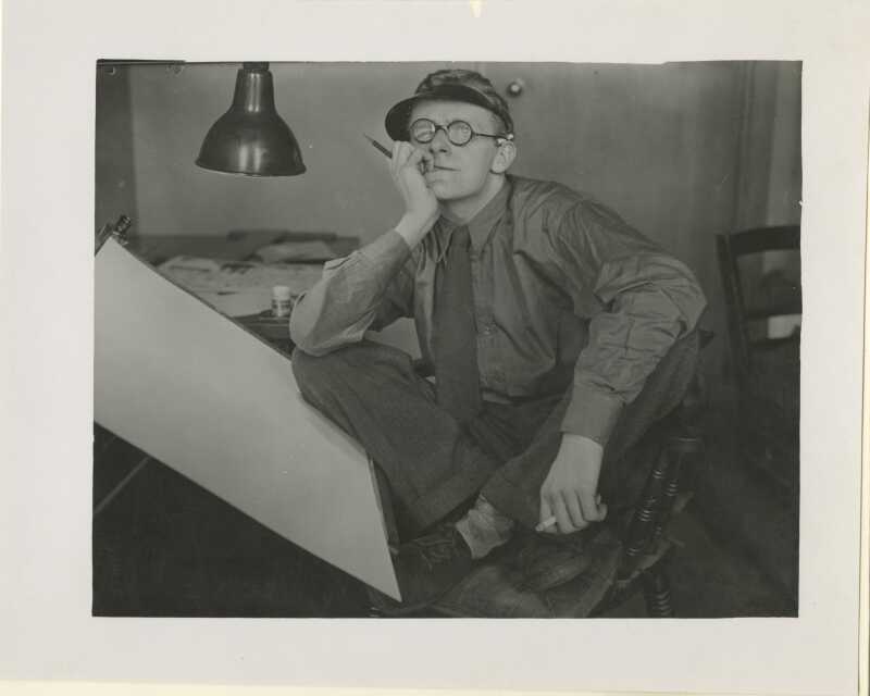 Black and white photo of Giles sitting cross-legged on a chair by his easel