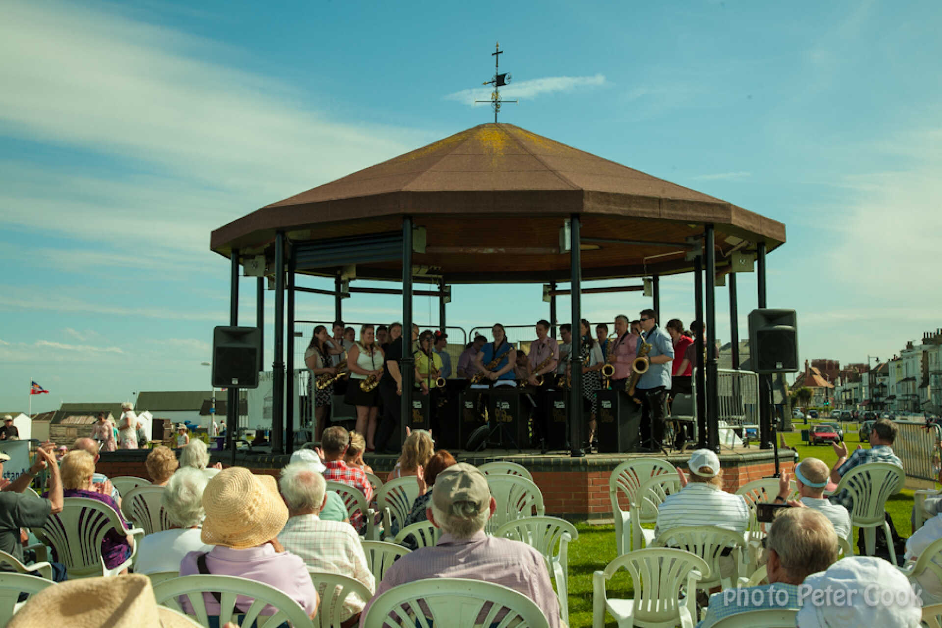 Audience seated around an outdoor bandstand against a blue summer sky