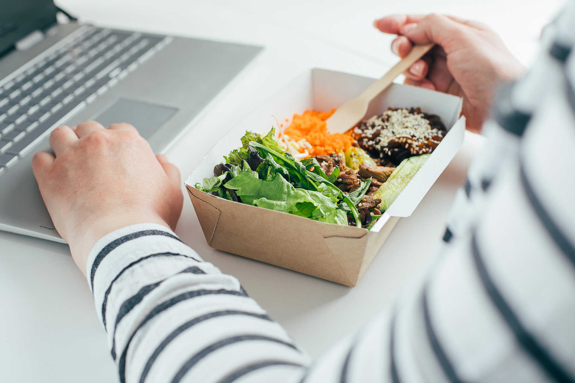 An image of someone typing at a laptop with one hand and eating a healthy, boxed salad with the other.