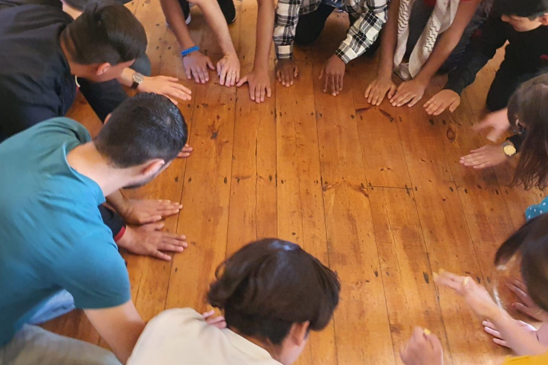 Group of people kneeling in circle on wooden floor with hands placed flat on floor in front of them.