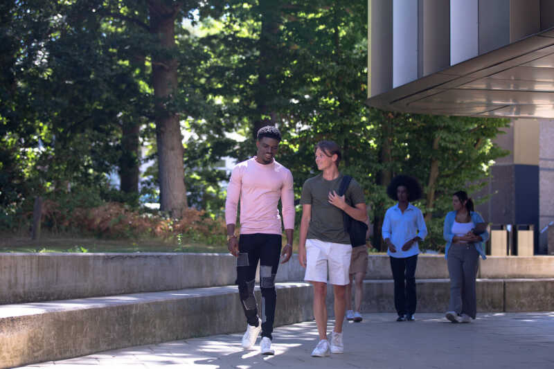 Two pairs of Kent students walking in conversation in dappled shade on concrete path next to contemporary building.