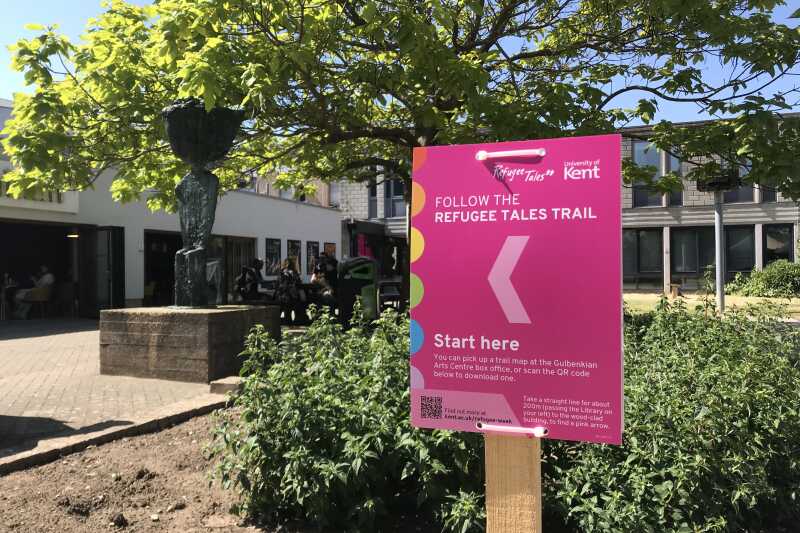 Pink Refugee Tales Trail sign outside the Gulbenkian Cafe in summer.