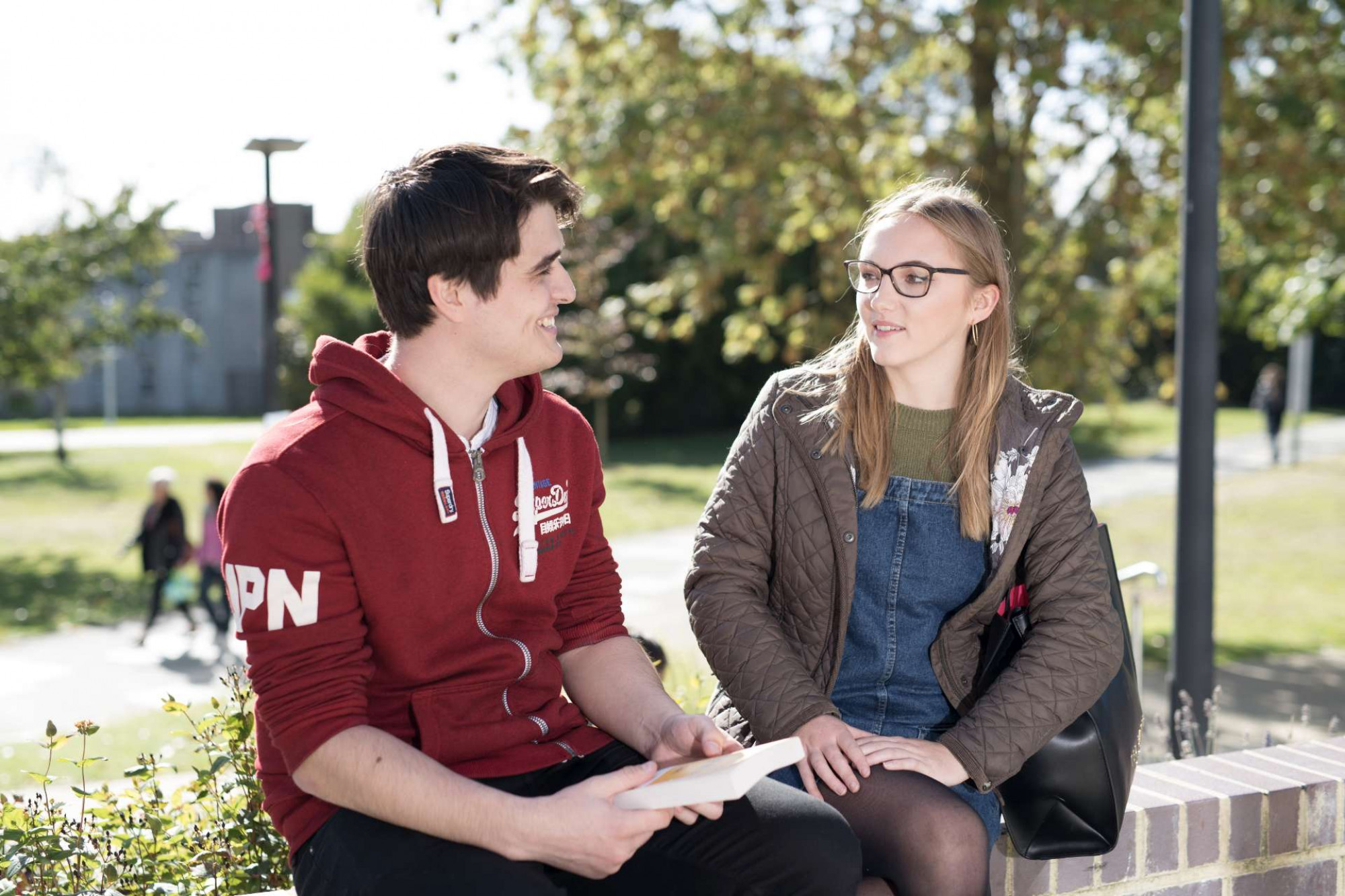 Male and female University of Kent students enjoying a chat outdoors on a sunny day, campus in the background.