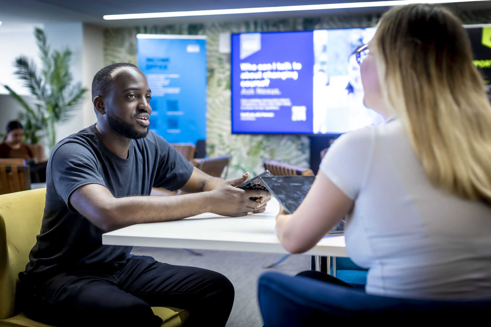 photo of young black man at a welcome desk speaking to a woman who is holding a tablet; display screen in background
