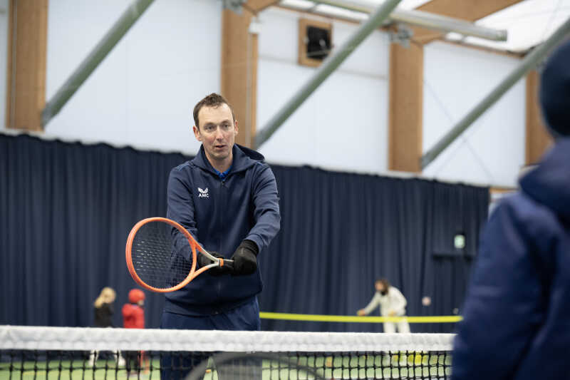 Lead coach Nick Skelton holding a tennis racket and giving pointers on correct technique.