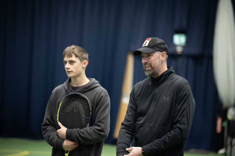 Two tennis players watching and listening to advice from a qualified coach.