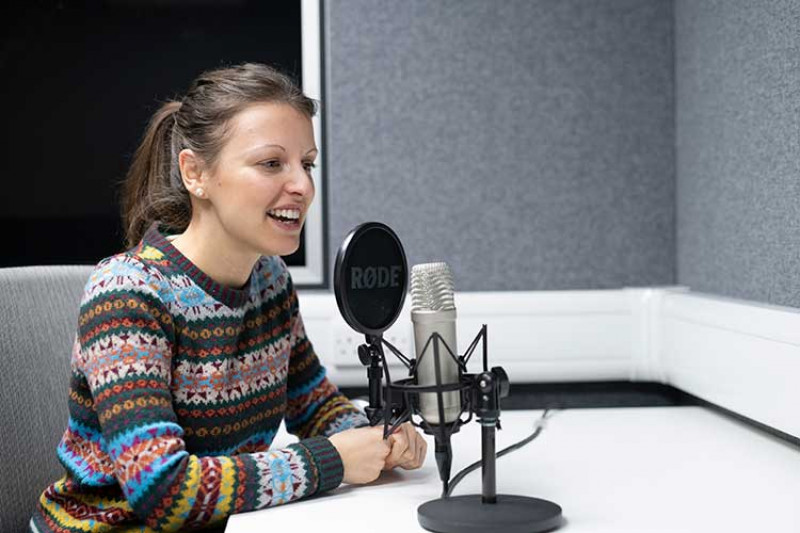 Woman in multi-coloured jumper speaking into a circular microphone in a studio