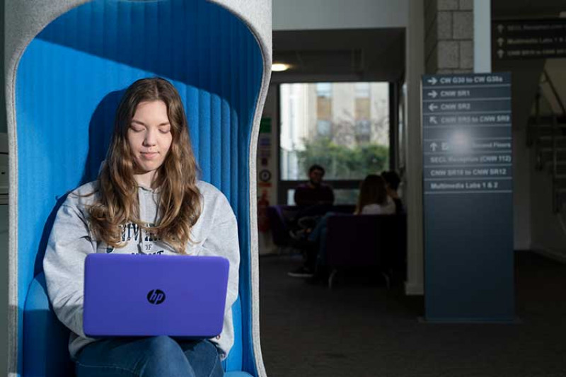Student with long brown hair, grey sweatshirt, sitting in a blue chair and using a purple laptop