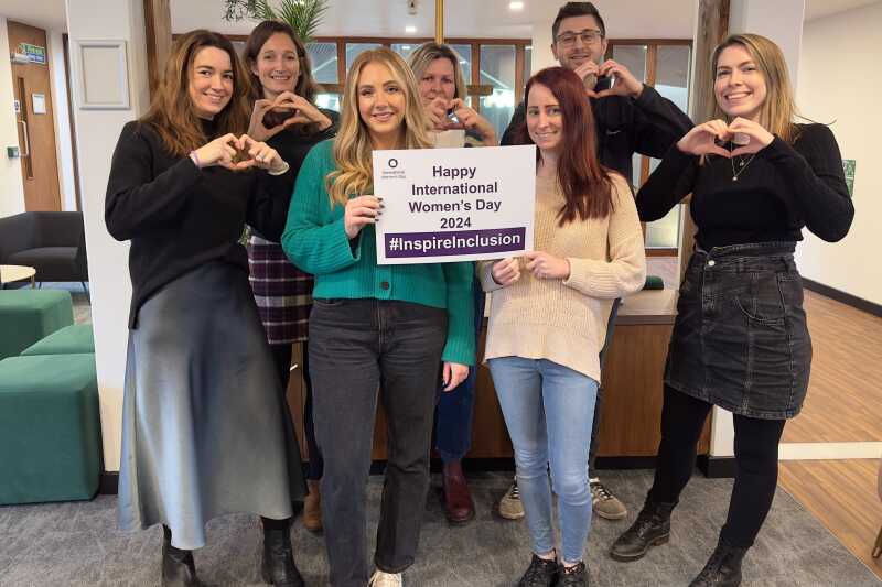 Communications team in the Rutherford lounge smiling and holding up an International Women's Day pledge card and a heart pose