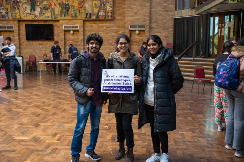 Three students in the Rutherford Dining Hall holding up an International Women's Day pledge cards