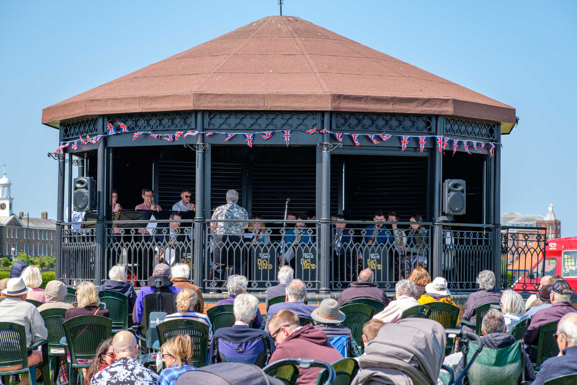 Musicians playing at a bandstand on a greensward, audience looking on