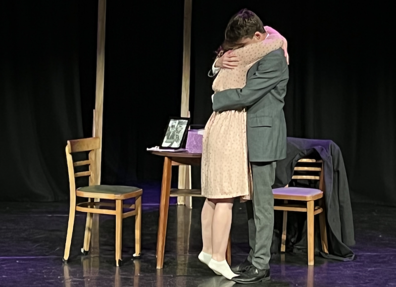 male and female embrace on a stage with just 2 wooden chairs and a table