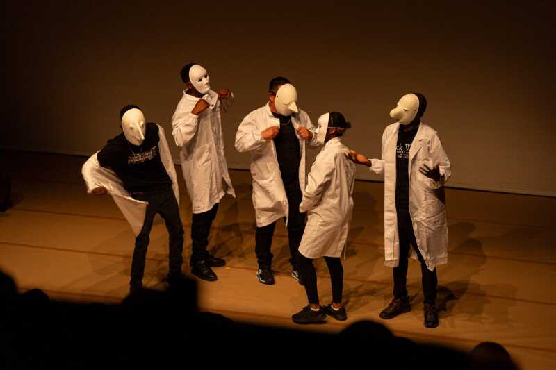 Theatre performers in white masks and lab coats