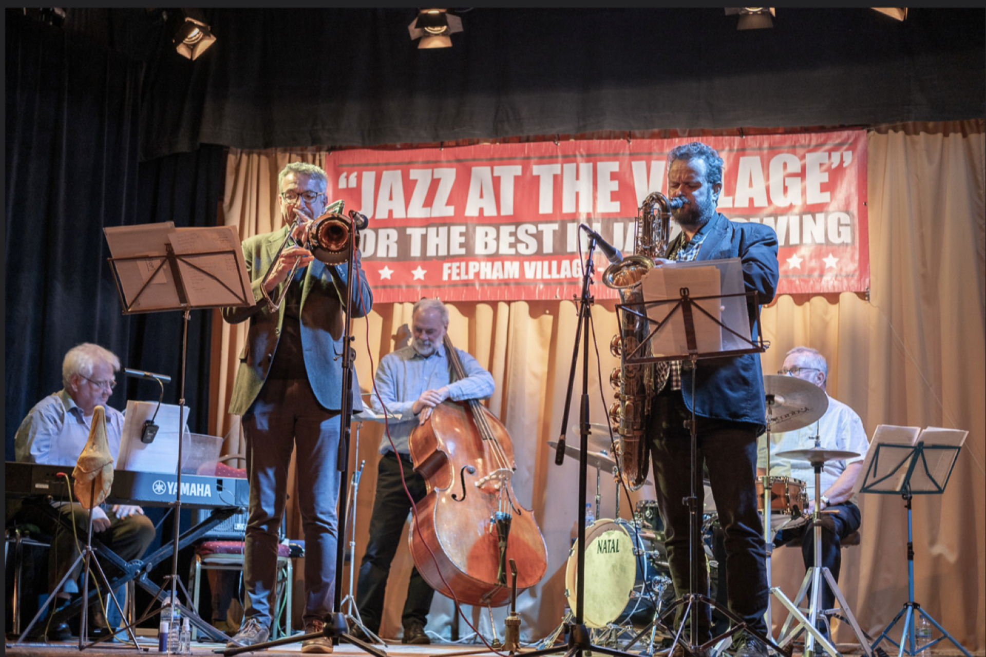 Group of six musicians playing on a stage, in front of a banner reading Jazz at the Village