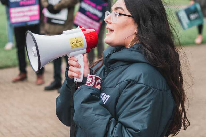 Image shows a young women speaking into a loudspeaker in front of a crowd