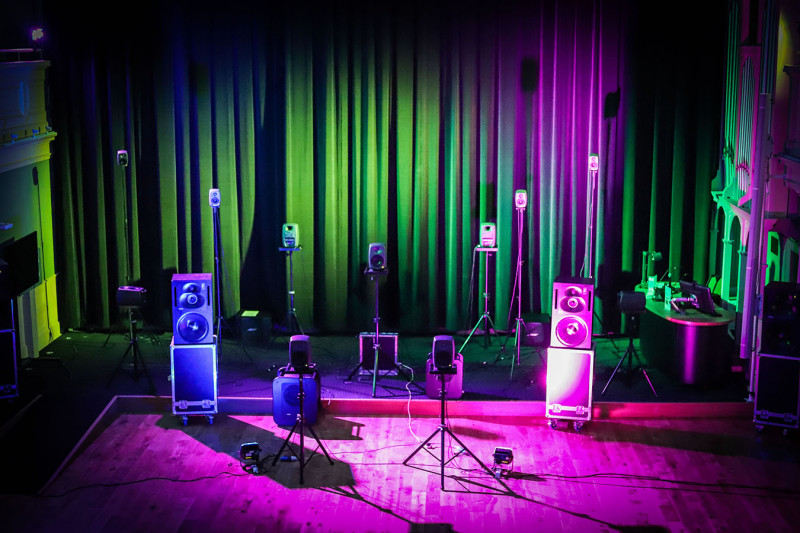 The Music and Audio Arts Sound Theatre set up in front of a closed curtain. Pink and green lights shine on it.