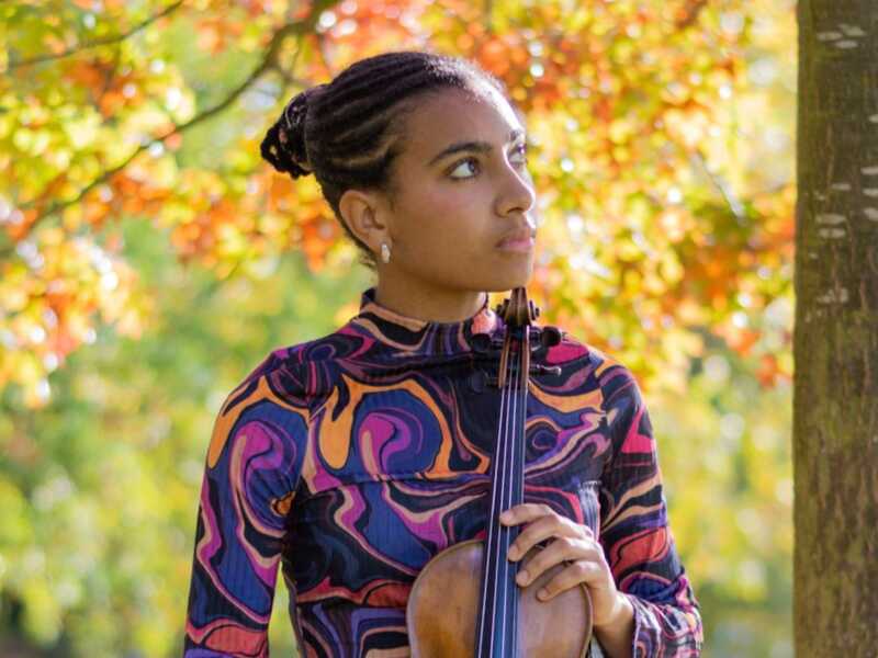 Female holding violin and looking over her left shoulder against a backdrop of tres