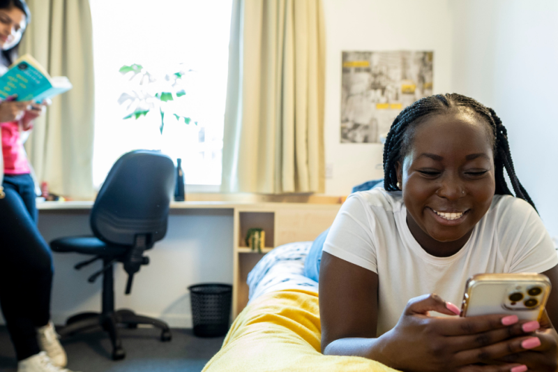 Image shows a young woman lying on a bed smiling as she plays with her phone