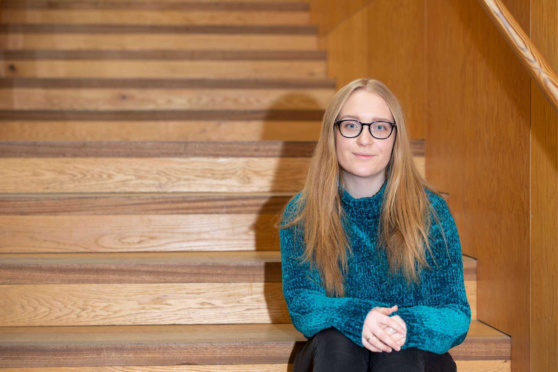 Shauna sits on a wooden staircase, wearing a teal jumper and black glasses.