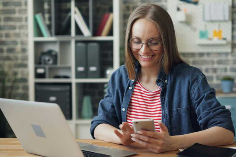 Lady sat at a table on her laptop viewing her mobile phone