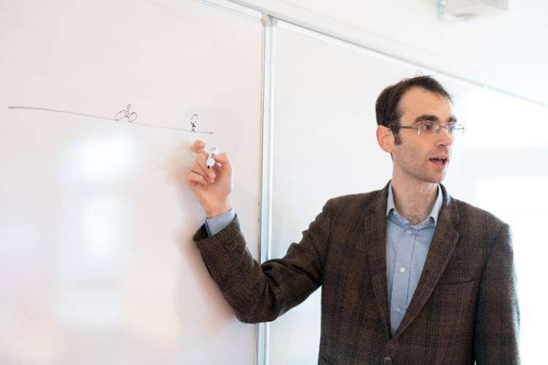 Dr Graeme Forbes, wearing glasses, a blue shirt and brown check jacket teaching at a white board.