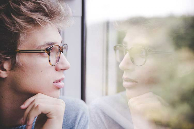 Young man with short brown hair and glasses leaning against a window and looking out of it.