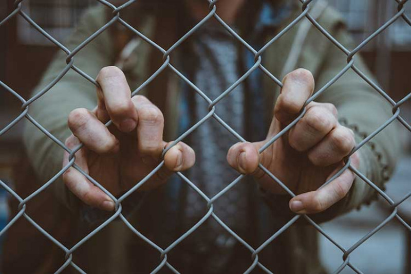 Man standing behind a wire fence, clutching the fence with both hands.