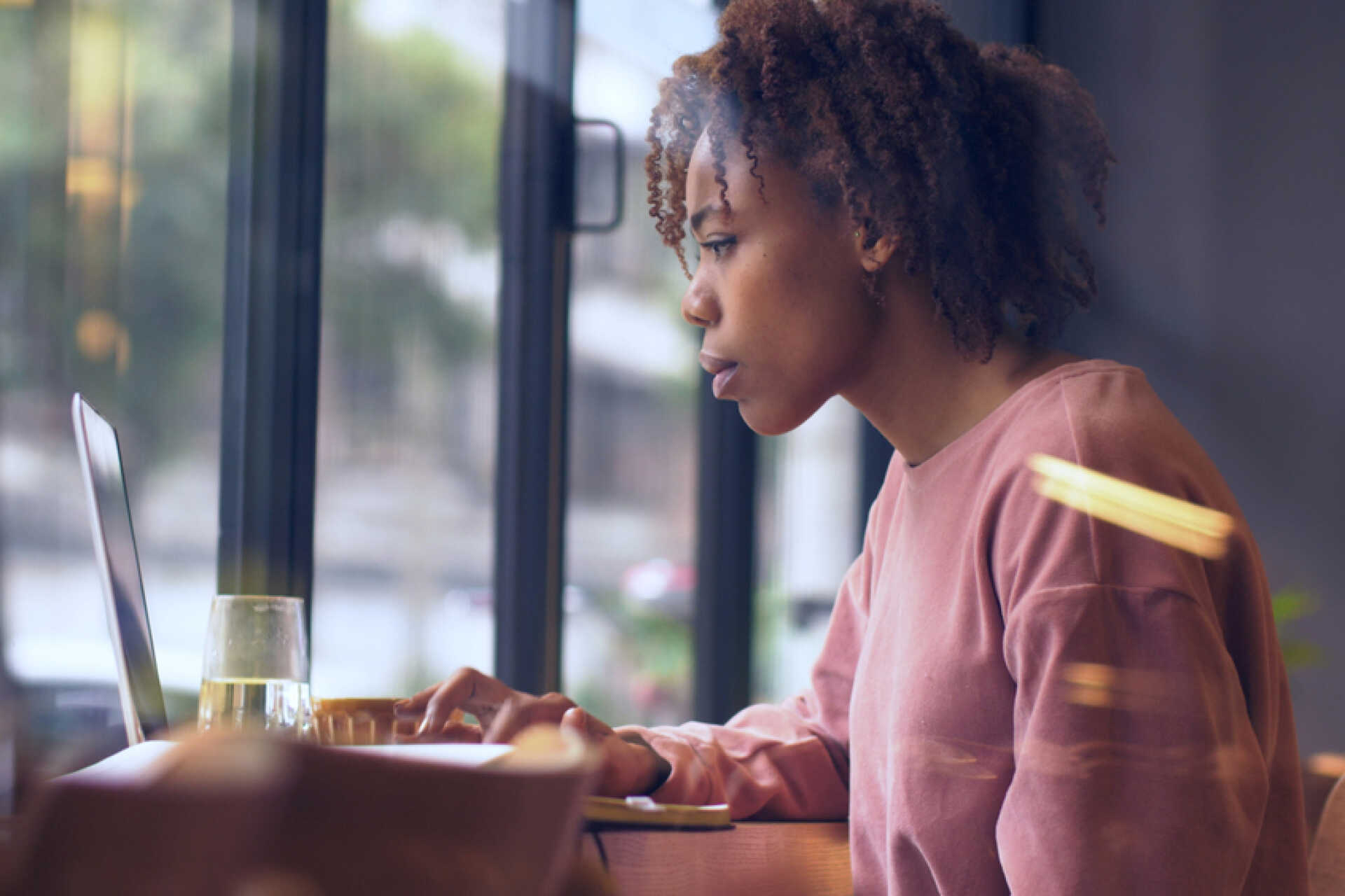 Side view of one young African woman sitting with a laptop by the window surfing the internet
