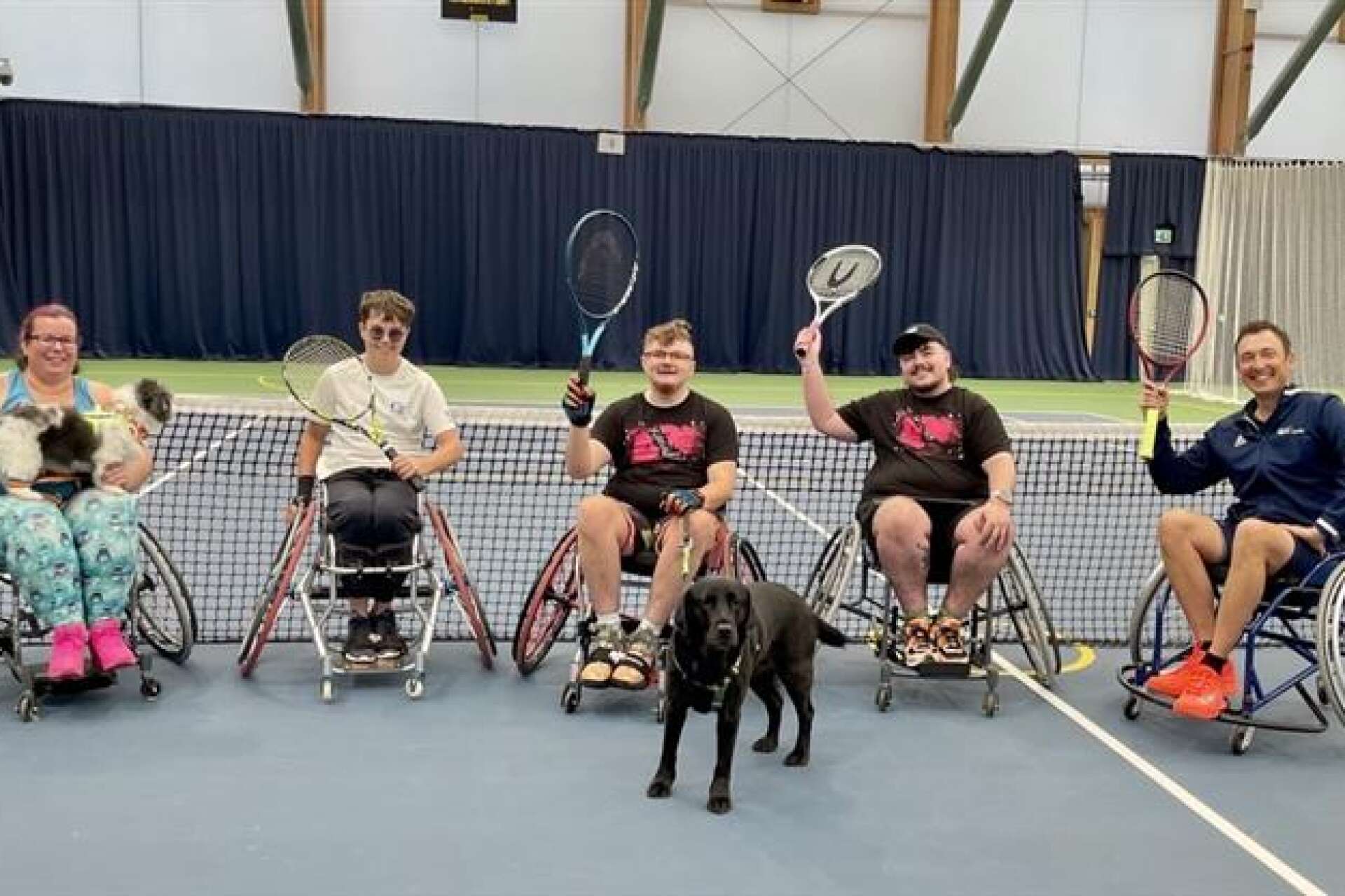 Group of people in tennis wheelchairs all raising their tennis rackets as they pose for a photo, with an assistance dog