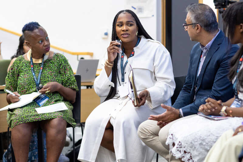 Image shows a woman talking into a microphone and people listening