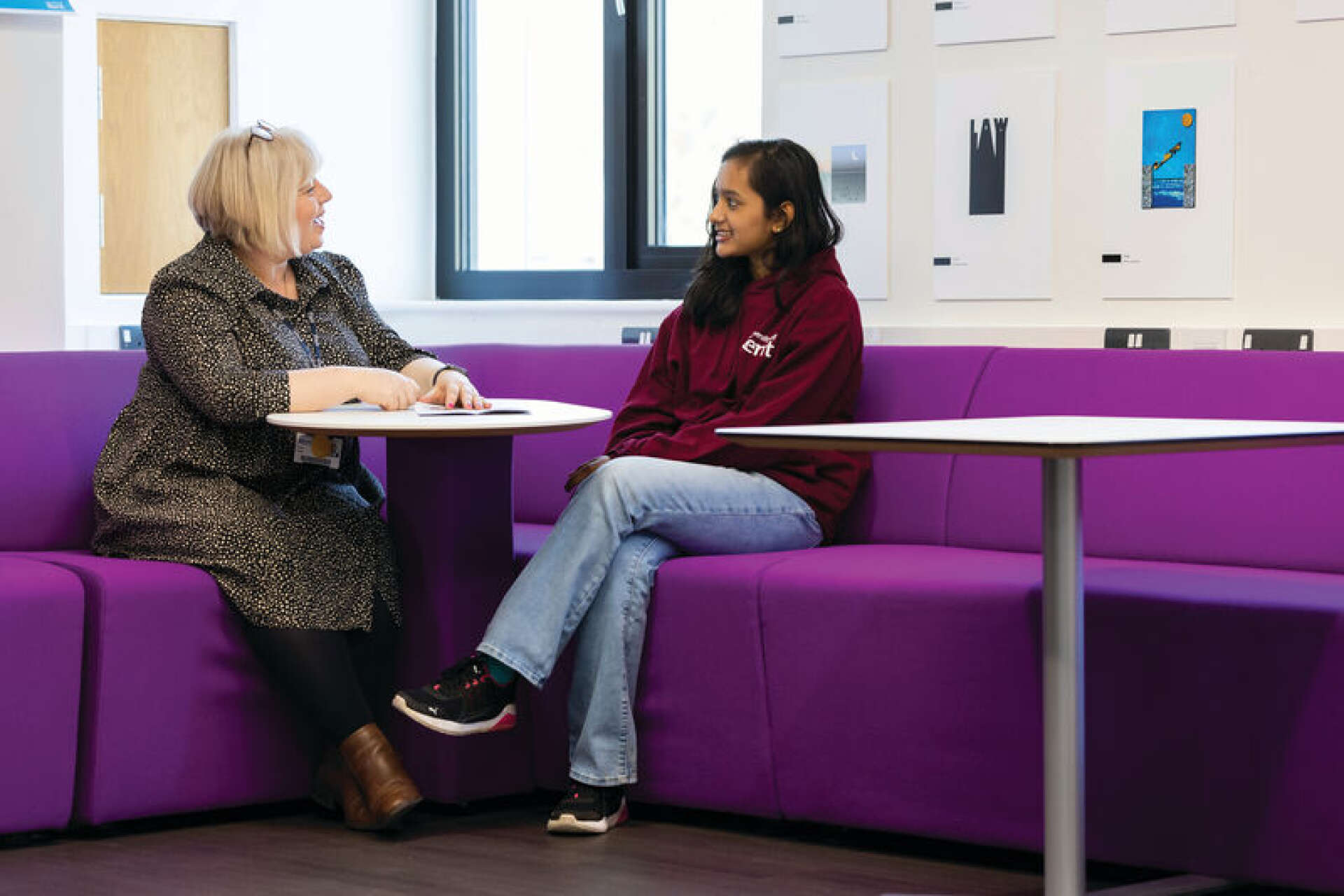 Staff member talking with a University of Kent student seated on purple seating in a campus common area.
