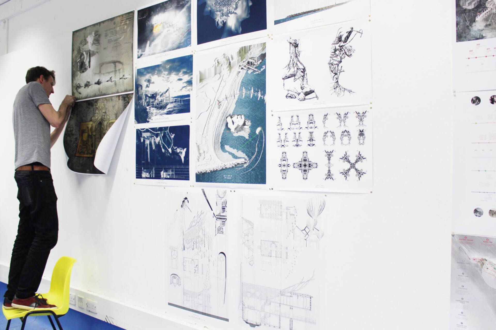 A student standing on a chair pinning his artwork to display on a wall with other pieces.