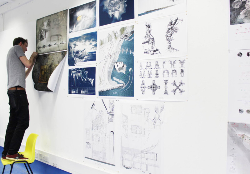 A student standing on a chair pinning his artwork to display on a wall with other pieces.